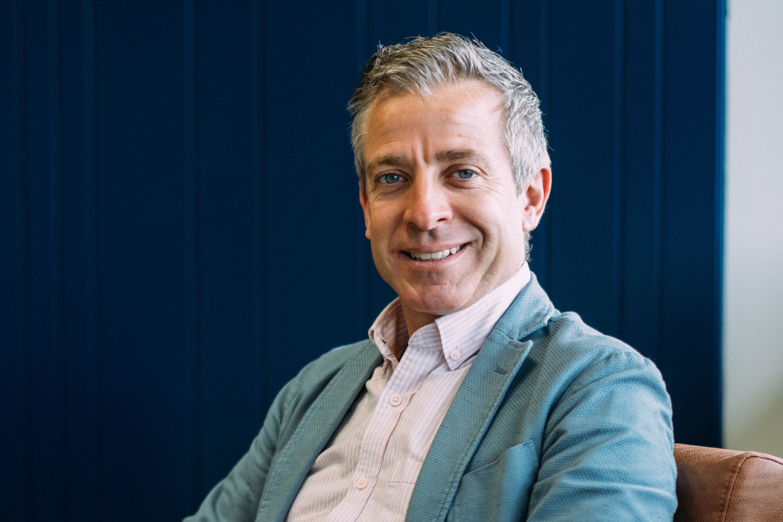 Photo of Dave Payne sitting down smiling into the camera wearing a white shirt under an eggshell blue suit jacket