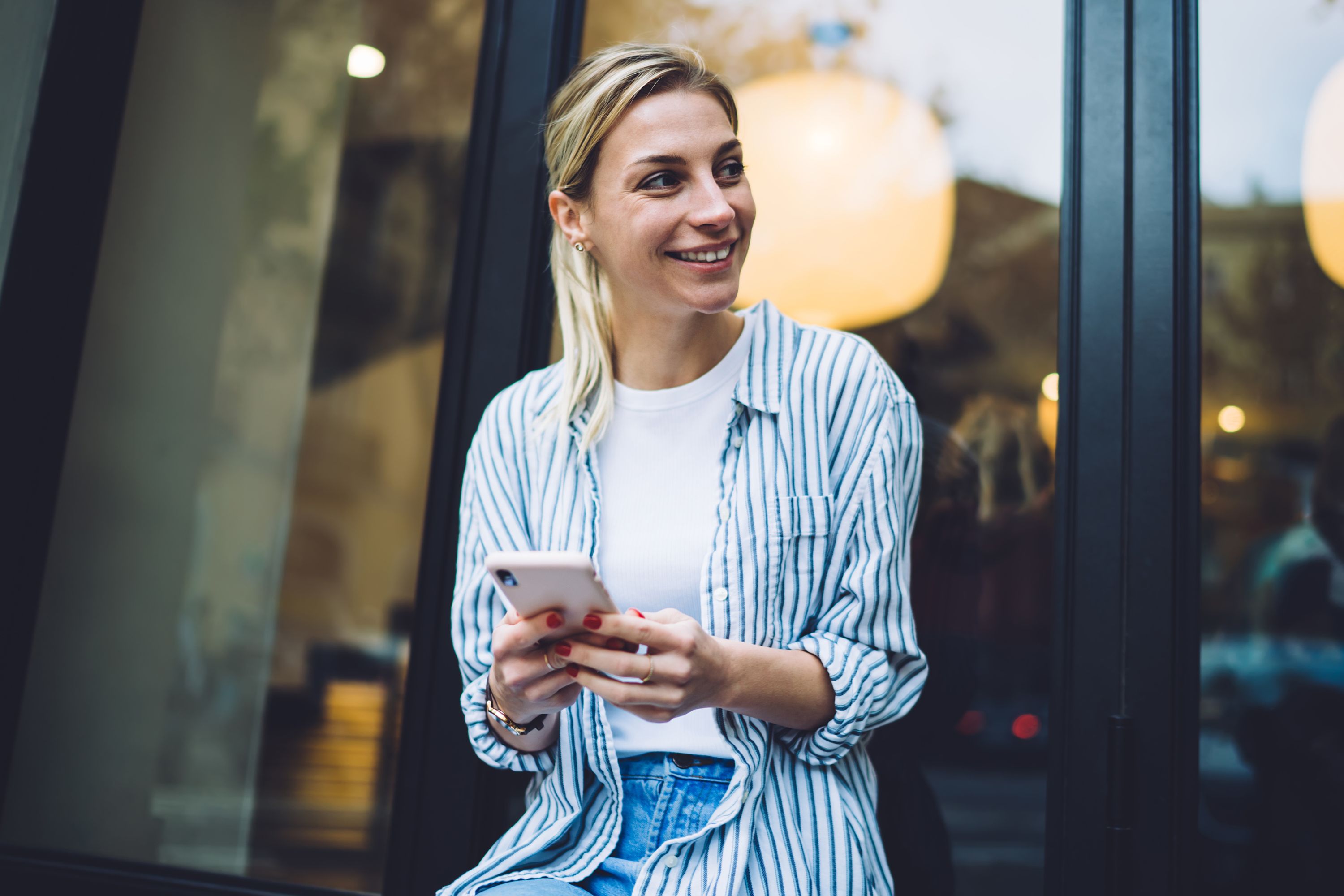 Photograph of a woman wearing awhite and blue striped shirt over a white t-shirt looking off to the side smiling and holding a mobile phone