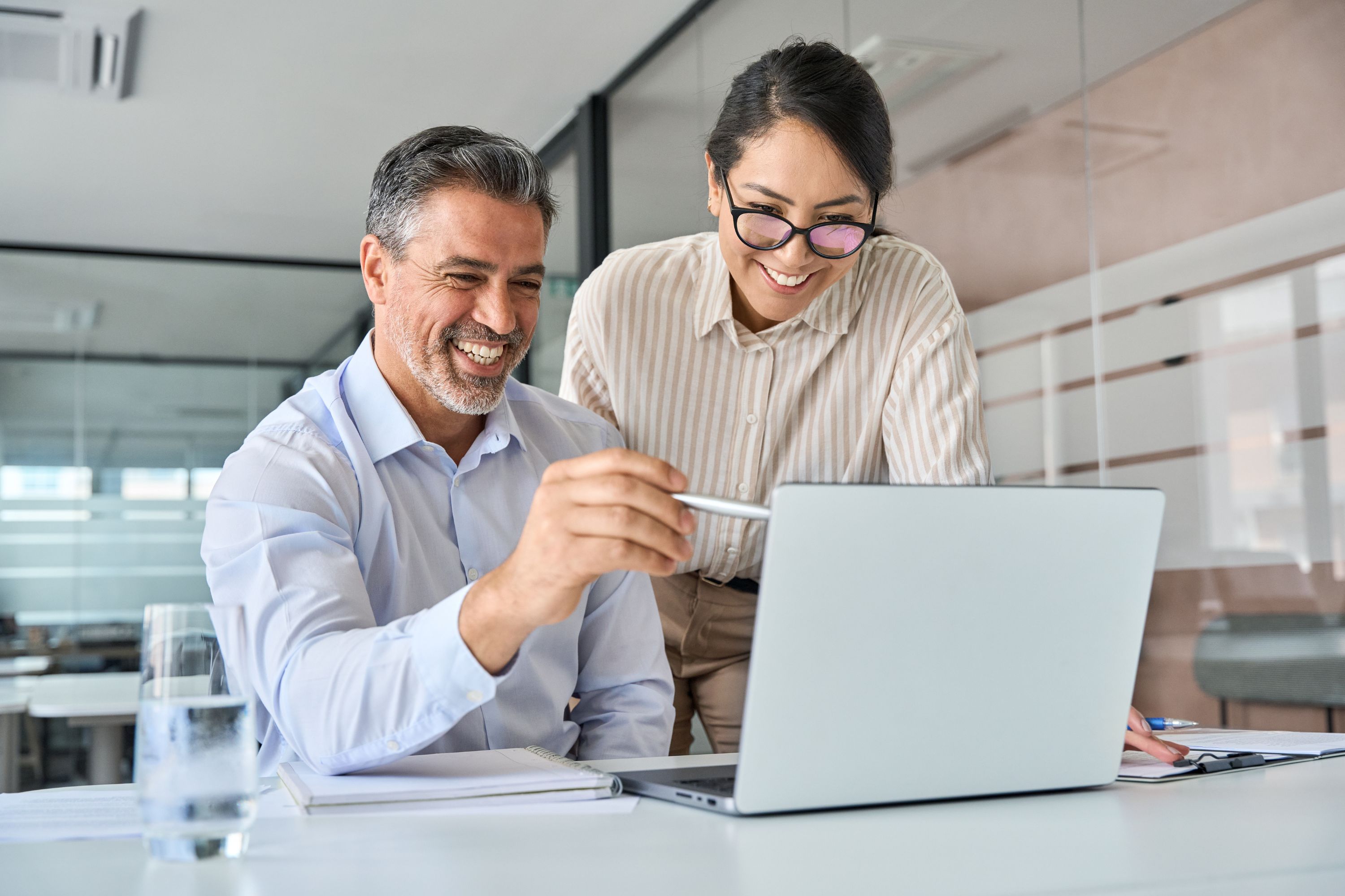 Two colleagues man and women in office meeting room looking at computer and smiling, bright atmosphere