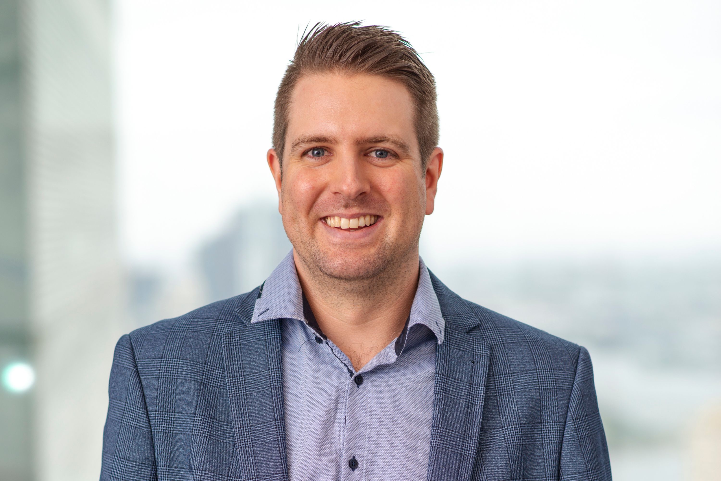 Portrait of Jacob Rothemund smiling and wearing suit in office setting, standing against blurred background of cityscape outside of window