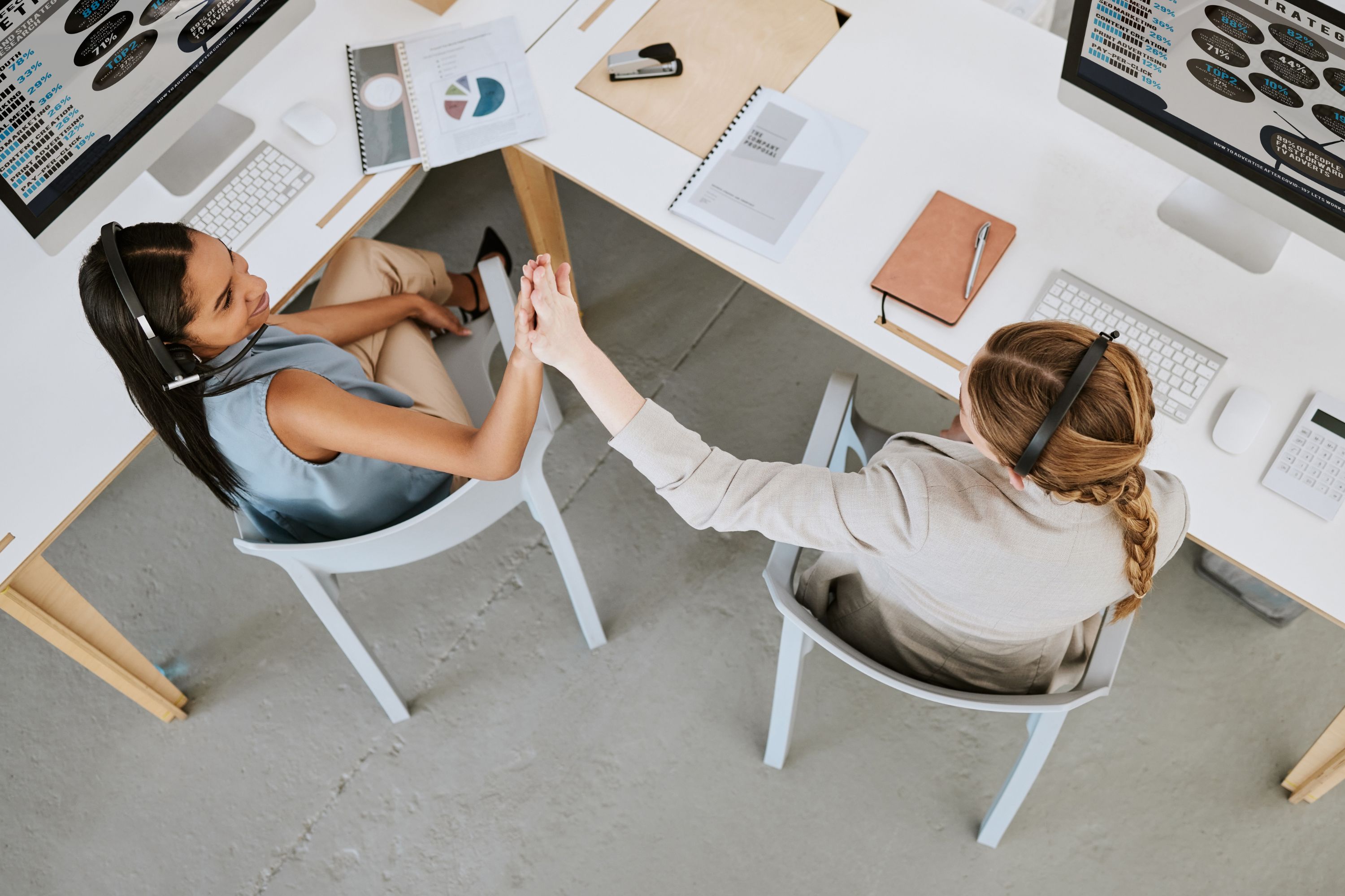 Birdseye view of two women sitting at desks giving each other a high five.