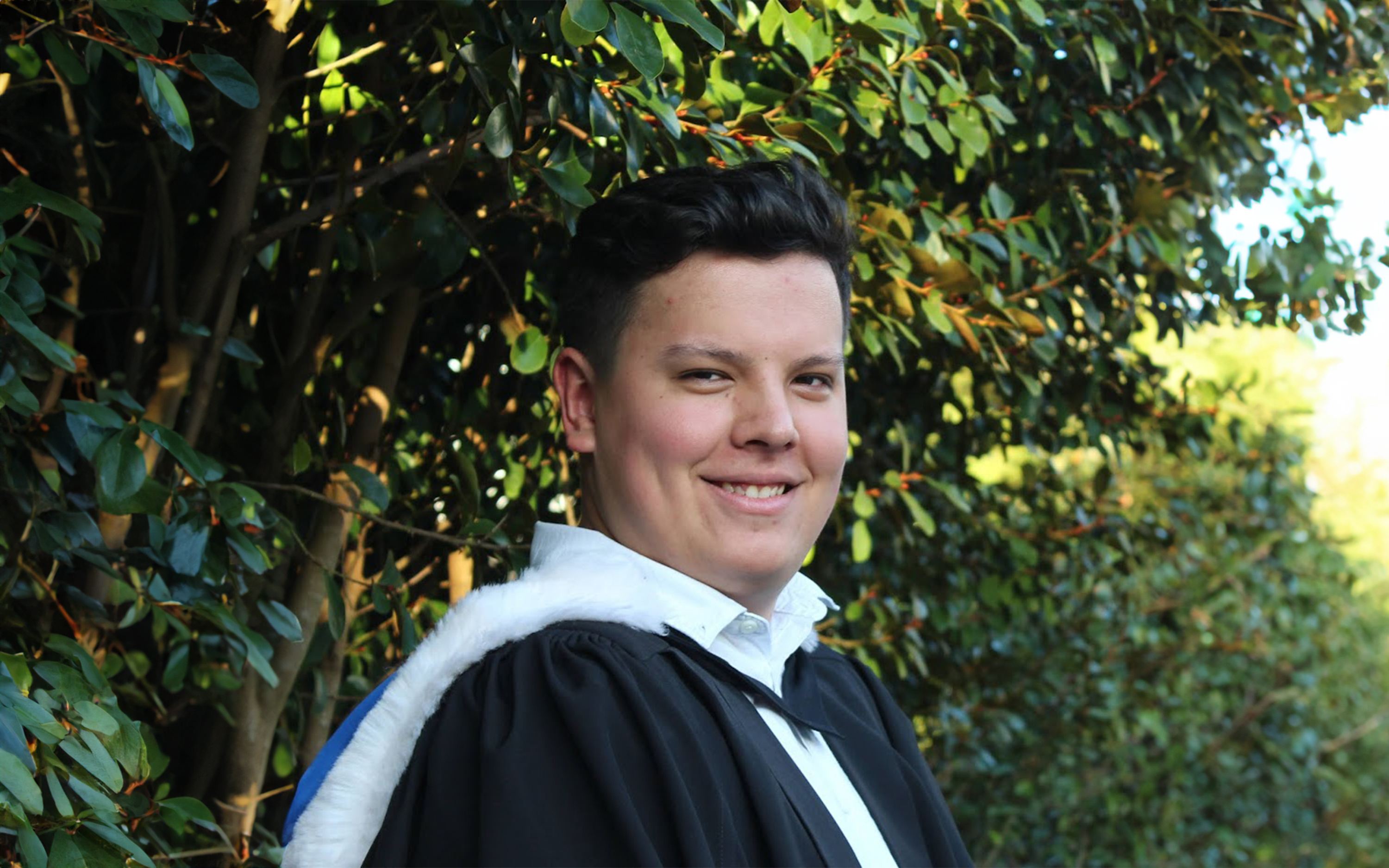 Photograph of Baxter Kirikiri smiling directly at the camera wearing a graduate gown.