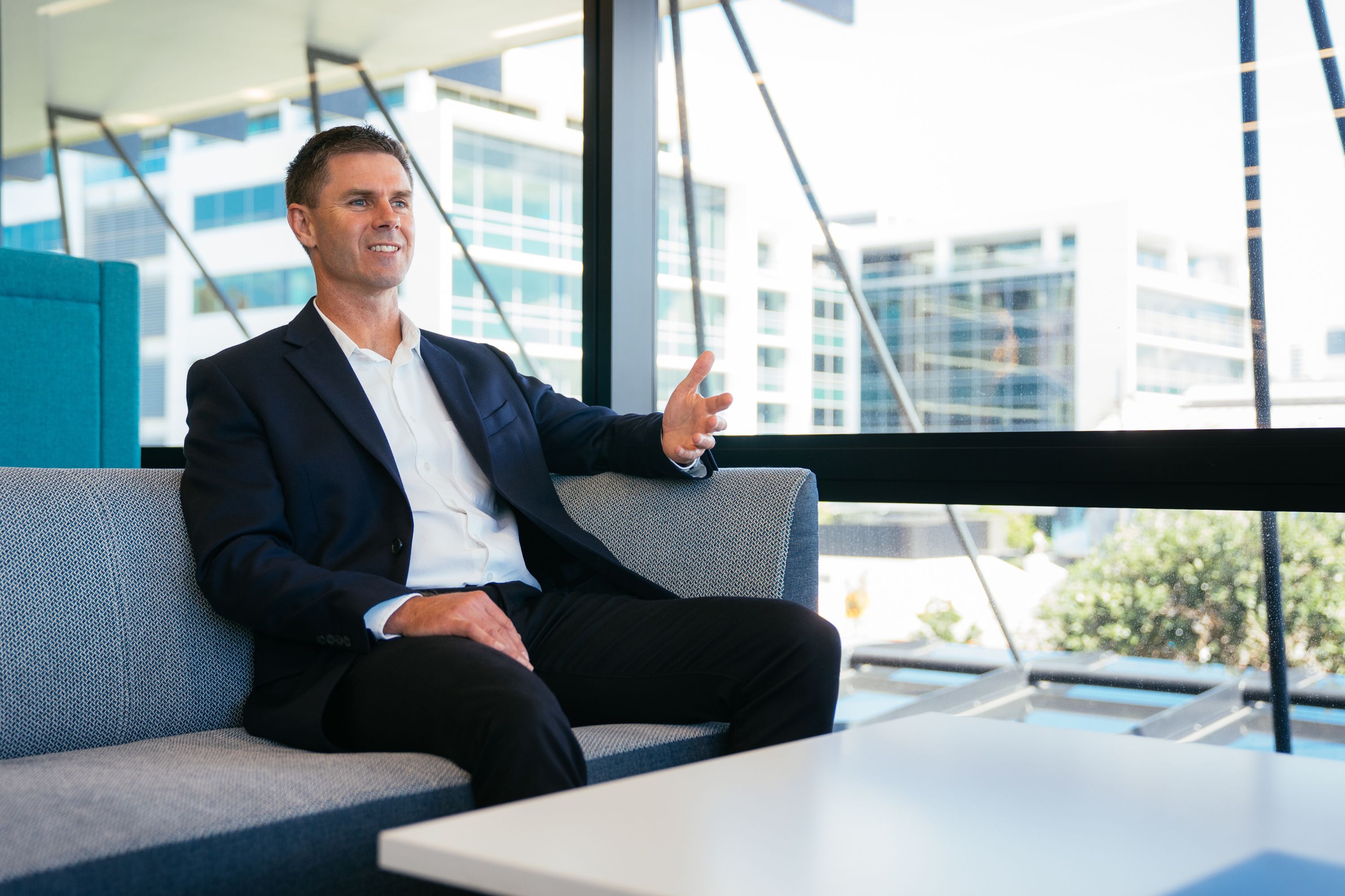 Photograph of Peter Nelson sitting on a long grey sofa next to a large window