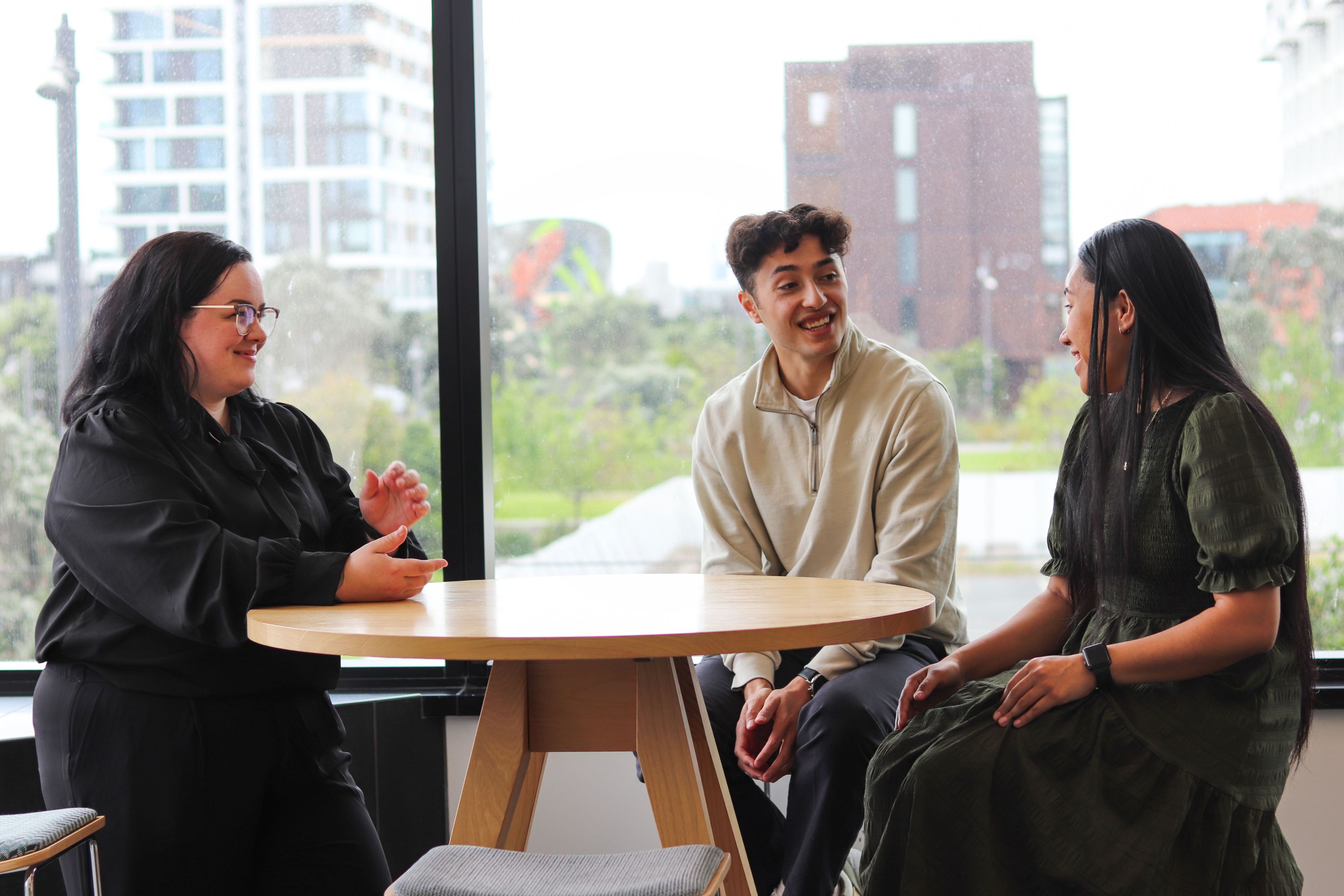 Photograph of three young people talking around a table in front of a large window.